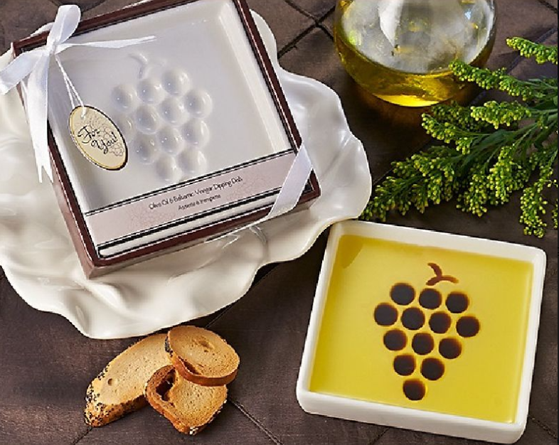 Small white squared dip plate with a grape design, surrounded by greenery, a golden glass of water on ice, the dip plate filled with olive oil and three bread toasts.