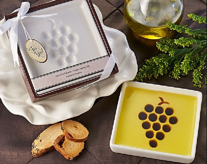 Small white squared dip plate with a grape design, surrounded by greenery, a golden glass of water on ice, the dip plate filled with olive oil and three bread toasts.