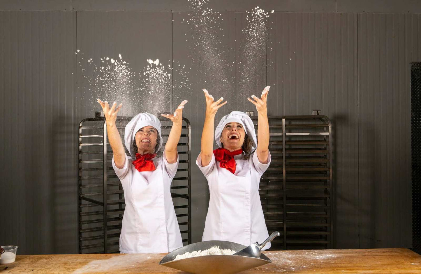 Two women in chef attire, throwing flour into the air in a kitchen setting.