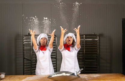 Two women in chef attire, throwing flour into the air in a kitchen setting.