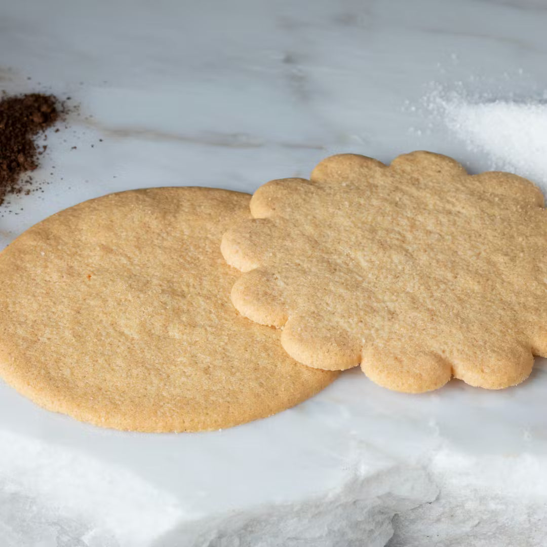 Two sugar cookies, one plain and one with a scalloped edge, on a marble surface.